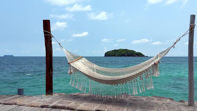 Hammock Swaying in the Breeze by Turquoise Sea. Empty hammock gently swaying in the breeze by turquoise sea with a small island in the background, peaceful seaside vacation and relaxation concept.