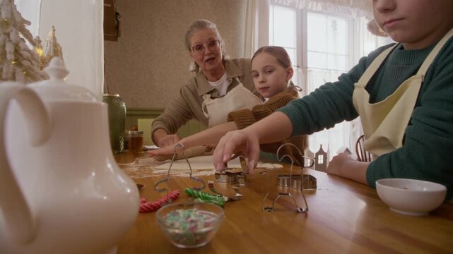 Senior Caucasian woman and children rolling dough and cutting holiday cookies at table. Clip suitable for Christmas holiday baking campaigns and intergenerational family lifestyle promotions