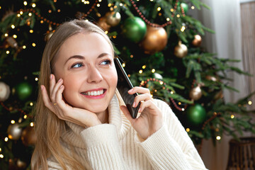 Close-up portrait of a happy young woman talking on the phone. Blurred Christmas tree lights bokeh in the background. Holiday greetings, calling loved ones, warm festive mood, copy space. © Anna Voloshyna