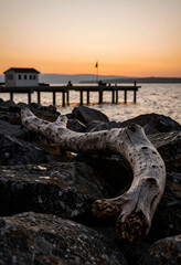 Fototapeta premium Serenity and mindfulness driftwood on rocky lakeshore at sunset with pier and calm water travel and nature escape peaceful coastal landscape