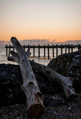 Fototapeta premium Serenity and mindfulness, driftwood on a rocky beach at sunset with a wooden pier in the background for travel and coastal nature relaxation concepts