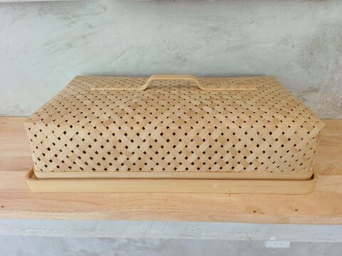 A traditional handcrafted bamboo woven food cover placed on a light-colored wooden shelf against a textured concrete wall.