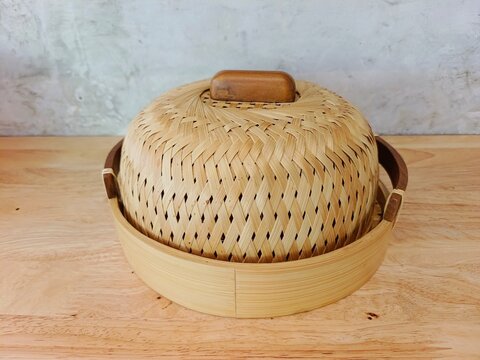A handmade woven bamboo food cover with a wooden handle placed on a light wood surface against a textured concrete wall background.