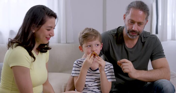 Young son bites carrot stick between Asian mother and Caucasian father on sofa as parents watch and talk, creating healthy snack time, supportive parenting, and joyful family connection at home.
