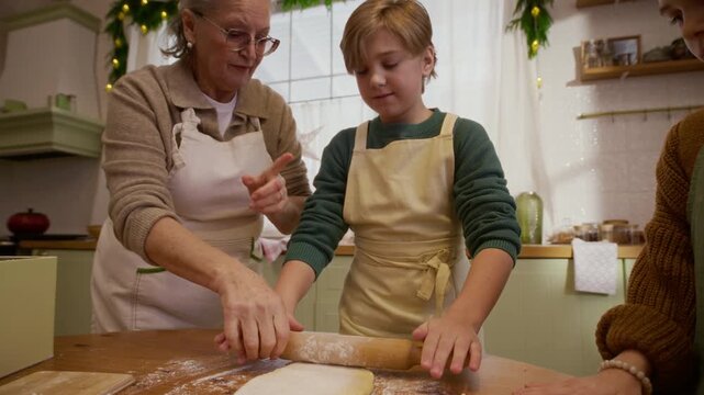 Senior Caucasian grandmother helping children roll pastry dough on table with rolling pin, zooming out. Clip suitable for holiday cooking ads and family lifestyle campaigns