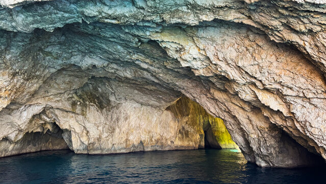Interior of a large limestone sea cave with layered rock walls, warm golden and green light at the exit and calm deep blue water, Paxos, Ionian Islands, Greece