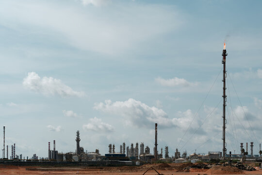 Tall industrial gas flare stack with guy wires burning off excess hydrocarbons at large crude oil refinery and petrochemical processing plant with fractionating columns under cloudy sky.