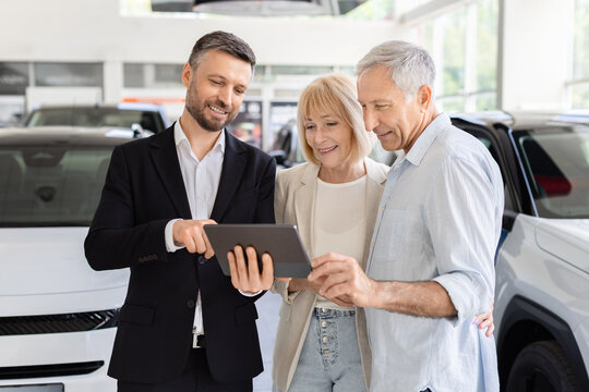 Car salesperson showing information on tablet to senior couple in dealership showroom as they review vehicle features and options before deciding on a purchase