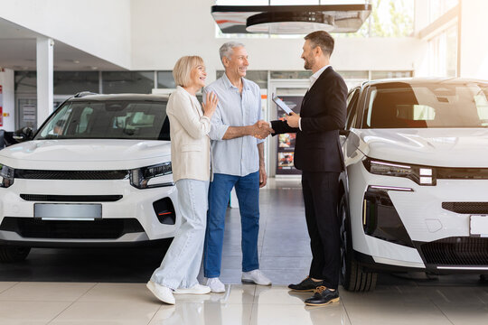 Senior couple shaking hands with car salesman between two new vehicles, sealing deal at modern dealership showroom