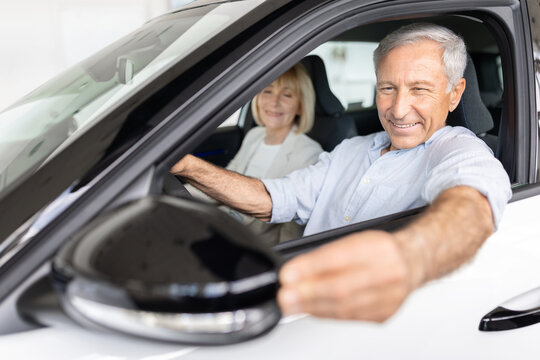 Cheerful senior man sits behind wheel of new car reaching toward side mirror at a dealership showroom, wife smiling in passenger seat beside him