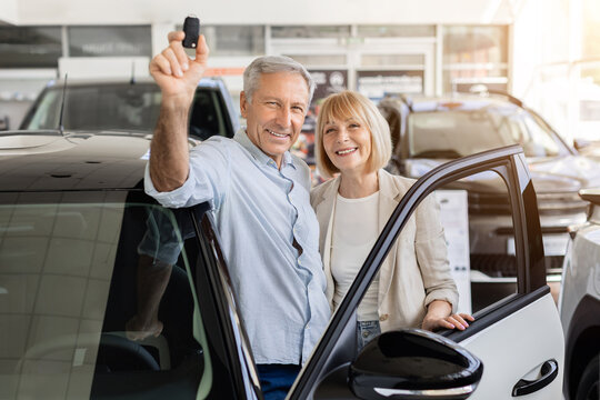 Cheerful senior couple standing by open car door at dealership, raising new vehicle key, elderly spouses celebrating fresh automobile purchase