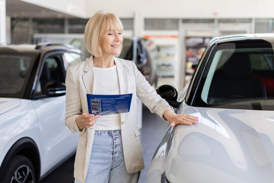 Senior Caucasian woman holding vehicle brochure and touching car in dealership showroom while comparing features and considering options before making a purchase
