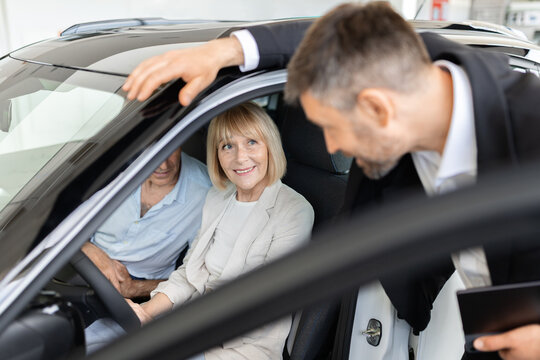 Senior Caucasian couple sitting inside new car while professional salesperson explains vehicle features through the open door during a dealership showroom consultation