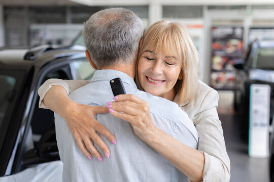 Happy senior Caucasian woman hugging her husband while holding car key inside a dealership showroom, celebrating together after purchasing a new vehicle and receiving ownership