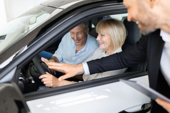 Car salesman pointing at steering wheel controls explaining features to smiling senior couple seated together inside new vehicle