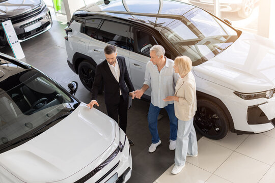 Senior couple comparing cars with professional salesperson in bright dealership showroom, discussing vehicle choices and exterior details before making a purchase decision