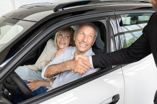 Senior man shakes salesmans hand through open car window while wife smiles beside him, elderly spouses finalizing new vehicle purchase deal