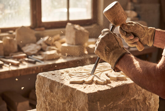 Artisan carving stone: A skilled artisan, wearing gloves, meticulously carves intricate patterns into a block of stone using a hammer and chisel in a sunlit workshop, dust flying.