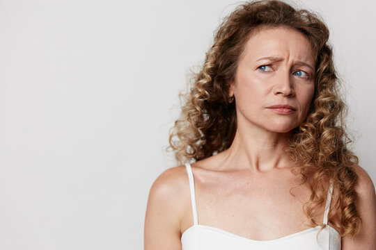 Authentic middle-aged woman with natural skin and curly hair looks away with worried expression. Unretouched portrait of mature female on a light background.