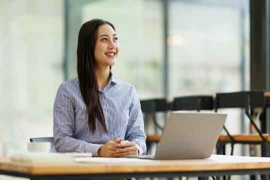 Asian businesswoman smiling, looking away from her laptop, and thinking, generating new ideas and future vision for business success while working in a bright office environment