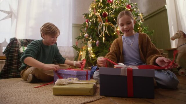 Caucasian boy and girl opening Christmas gift boxes near decorated tree in low angle shot. Footage fits holiday retail campaigns, toy commercials, and seasonal family promotions