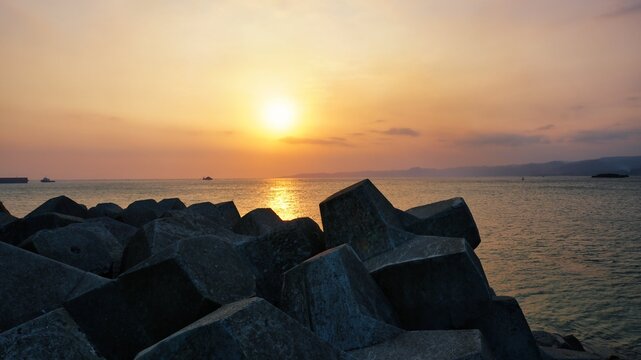 Stunning Golden Sunset Over the Calm Sea with Concrete Tetrapod Breakwater Rocks in the Foreground, Peaceful Coastal Landscape with Distant Mountains and Ships During Golden Hour at Twilight.