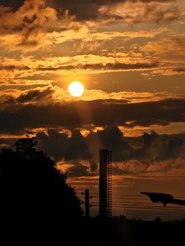 The sun sets, casting golden light through clouds. A smokestack silhouettes against the fiery sky.