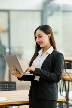 Asian businesswoman standing and smiling, working on her laptop in a contemporary office, focusing on tasks and leveraging digital tools for modern business operations