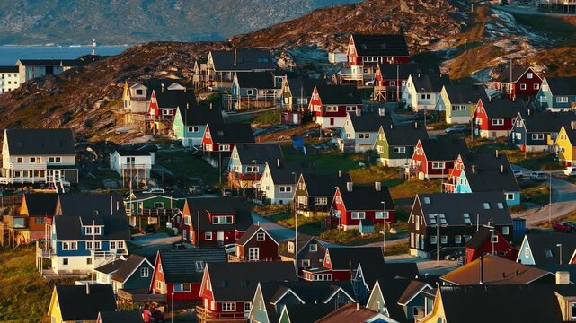 Cinematic drone footage of Nuuk, Greenland, captured during sunset, revealing colorful houses stretching along the Arctic coastline. Warm evening light reflects across the sea. 