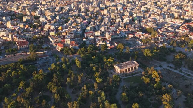 Golden hour sunlight illuminating the historic Temple of Hephaestus and the Ancient Agora, surrounded by the sprawling cityscape and dense green trees of downtown Athens, Greece from above