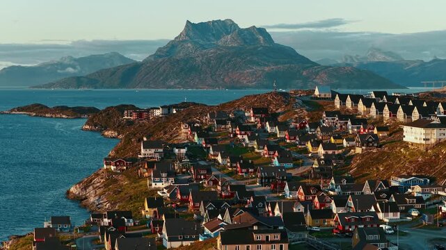 Cinematic drone footage of Nuuk, Greenland, captured during sunset, revealing colorful houses stretching along the Arctic coastline. Warm evening light reflects across the sea. 