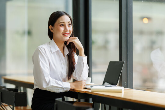 Asian businesswoman smiling, thinking about future success and opportunities, enjoying a moment of contemplation during her busy workday in a modern office environment
