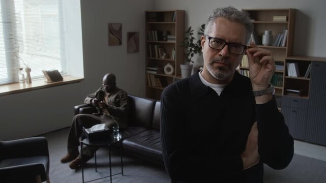 Portrait of middle aged Caucasian therapist looking at camera with crossed arms while African American patient sitting on couch. Clip suitable for mental health campaigns and therapy service promo