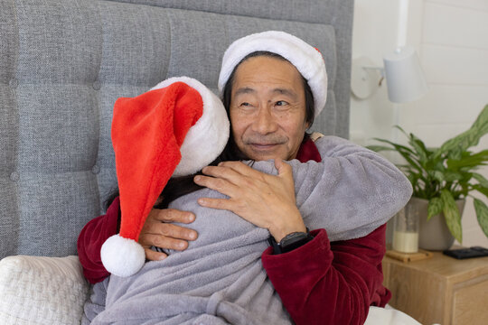 Senior Asian man and child hugging on bed, wearing Santa hats for christmas, gray headboard