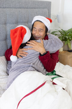 Senior Asian man hugging partner on bed on christmas morning, wearing red robe, Santa hats