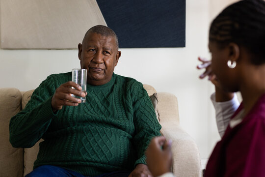 African American senior man holding glass of water, caregiver in scrubs gesturing on beige sofa