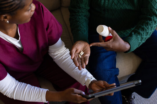 African American nurse and patient sitting at home checking tablet holding pill bottle and pills