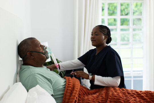 African American caregiver in scrubs adjusting oxygen mask and tubing, tending male patient in bed