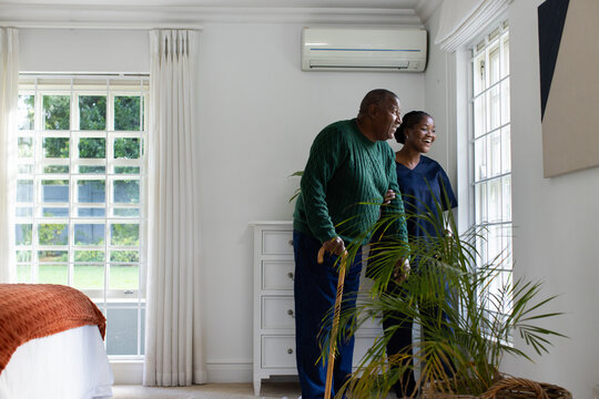 African American couple standing in bedroom senior man leaning on cane looking outside, copy space