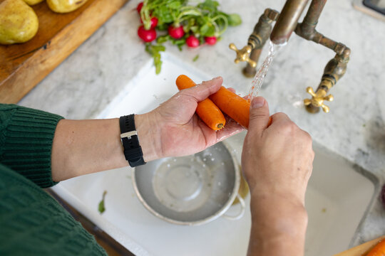Asian man washing carrots under brass faucet in kitchen sink, in green sweater and watch