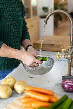 Mature Asian man washing peppers in colander under kitchen faucet wearing sweater, copy space