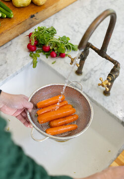Mature Asian man in green sleeves holding colander and rinsing six carrots under brass faucet