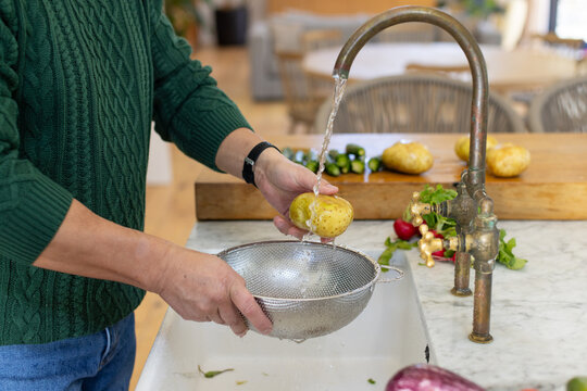 Asian man rinsing potato under curved brass faucet at marble sink holding stainless-steel colander