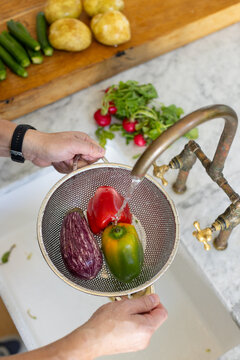 Asian man holding colander under running brass faucet, rinsing peppers and eggplant at kitchen sink