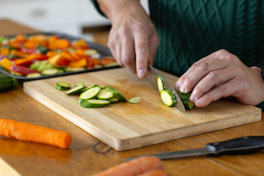 Mature man in dark green sweater slicing zucchini on wood board in kitchen, using chef's knife