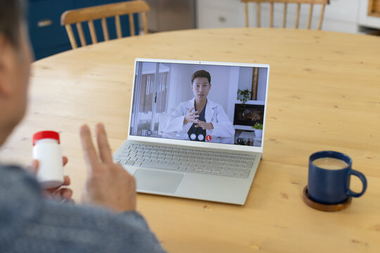 Senior patient holding pill bottle, consulting Asian doctor in lab coat on laptop at dining table