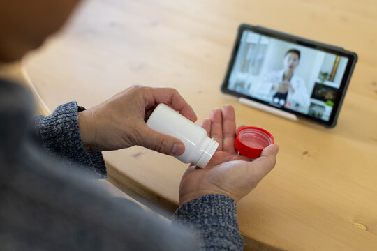 Male patient pouring pills from white-bottle, red-cap, blue-sweater at wood table, doctor on tablet
