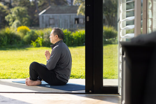 Mature Asian man sitting cross-legged on blue yoga mat on patio, wearing dark jacket, copy space