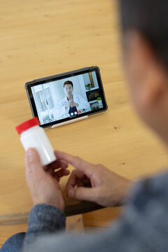 Mature man holding pill bottle at wooden table, viewing Asian doctor in lab coat on tablet