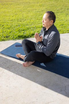 Senior Asian man meditating cross-legged on blue mat outdoors in sport jacket with phone
